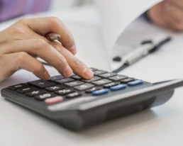 foto de manos sosteniendo lapiz y presionando botones de calculadora sobre documentos mini